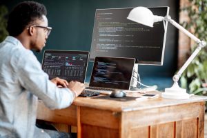 Young,African,Male,Programmer,Writing,Program,Code,Sitting,At,The Employee typing code from their home office with two laptops and a desktop